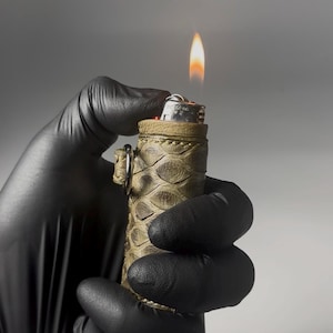 May include: A lit lighter with a flame, held in a gloved hand. The lighter is encased in a textured, olive-green snakeskin-patterned sleeve. A silver ring is attached to the side. The background is a gradient of grey.