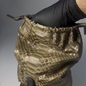 Puede incluir: Un bolso con cordón hecho de material texturizado con un patrón de reptil en tonos beige y marrón. El bolso está abierto, revelando el interior, y lo sostiene una mano enguantada. El diseño del bolso sugiere un enfoque en la textura y los patrones naturales.