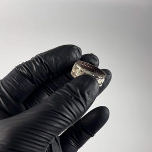May include: A silver ring with a textured design. The ring is held in a black gloved hand.