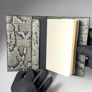 Peut inclure: Carnet ouvert avec une couverture en cuir vert et à motif peau de serpent. Le carnet a du papier crème ligné et un intérieur gris foncé. La couverture comporte des fentes pour cartes. Le carnet est tenu ouvert par des mains gantées.