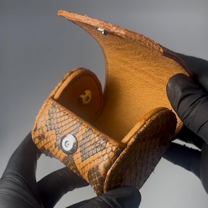 May include: Open, orange and brown snakeskin coin purse with a snap closure. The interior is a lighter shade of brown. The purse is held open by a person wearing black gloves.