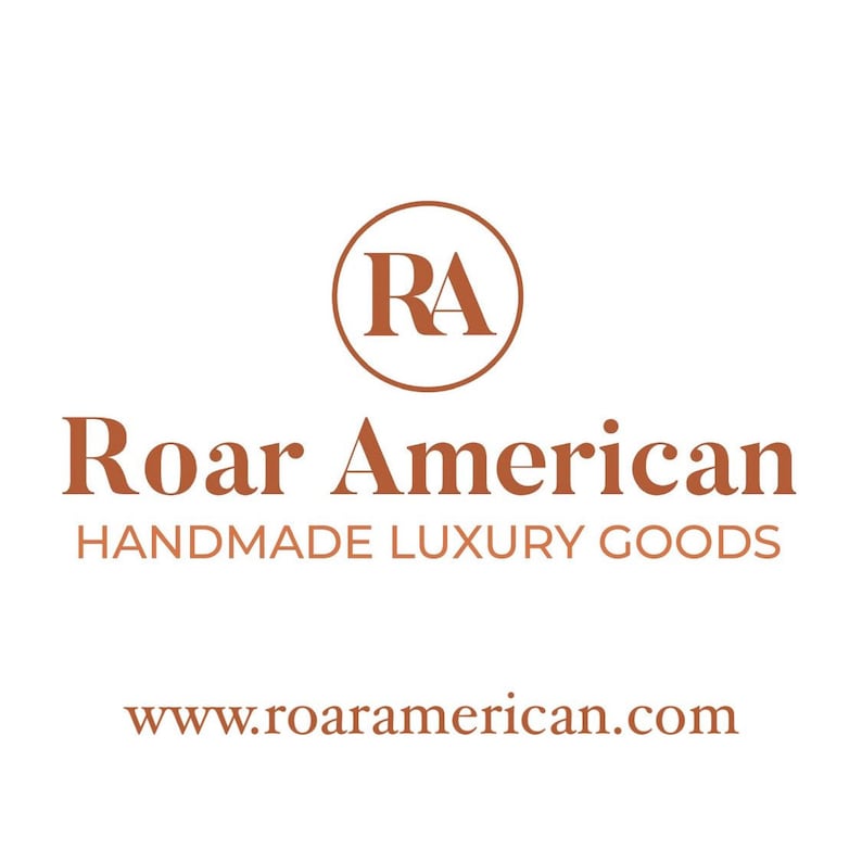 May include: Logo for Roar American, a company that makes handmade luxury goods. The logo features a circle with the letters "RA" inside, and the words "Roar American" and "Handmade Luxury Goods" below the circle. The website address, www.roaramerican.com, is displayed below the text.