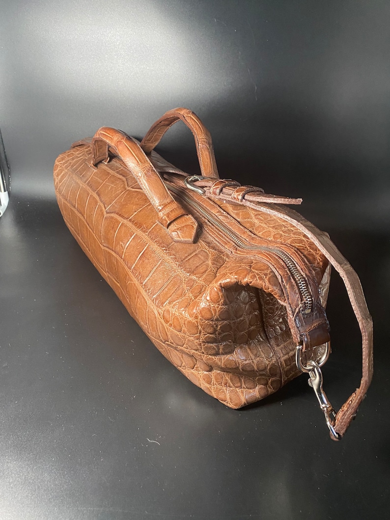 May include: A brown crocodile leather handbag with a top zipper closure and two handles. The bag has a detachable shoulder strap with a silver clasp.