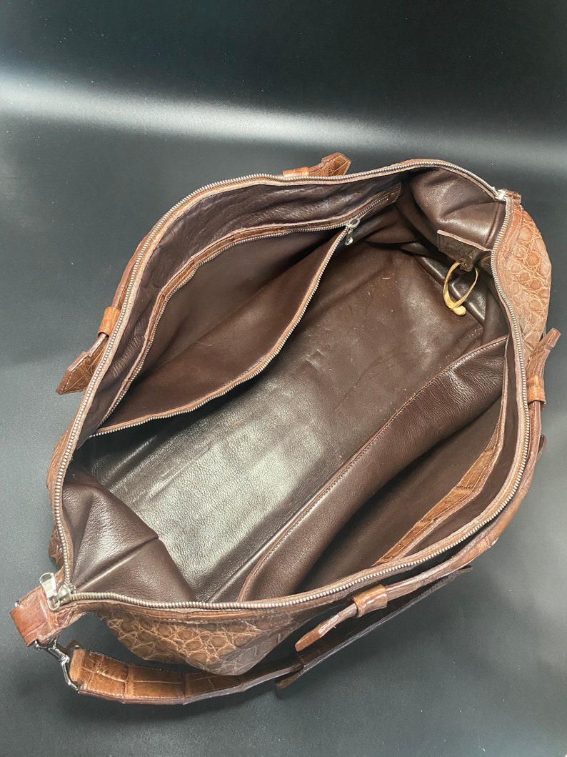 May include: A brown leather travel bag with a crocodile pattern. The bag is open and the inside is lined with brown fabric. The bag has a zipper closure and a shoulder strap.