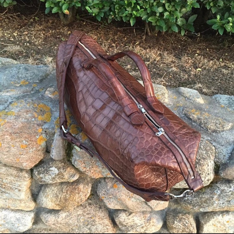 May include: A brown crocodile leather duffel bag with a zipper closure and a long strap. The bag is sitting on a pile of rocks.