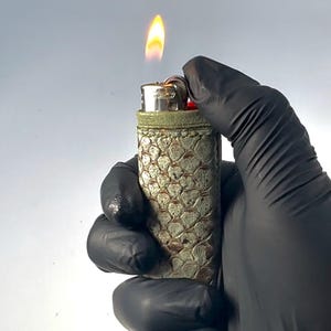 Peut inclure: Un briquet allumé avec une flamme. Le briquet présente un motif texturé en écailles dans des tons de vert et de beige. Le briquet est tenu par une main gantée.