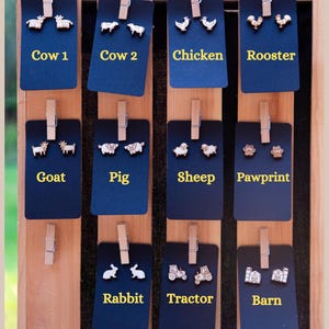 May include: A wooden display board with black cards featuring various farm animal and other themed earrings. The cards are labeled with the names of the animals and objects, including Cow 1, Cow 2, Chicken, Rooster, Goat, Pig, Sheep, Pawprint, Rabbit, Tractor, and Barn.