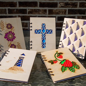 May include: A collection of wooden notebooks with laser-cut designs. The designs include a cross, flowers, a lighthouse, a strawberry, and a geometric pattern. Each notebook has a spiral binding and is made of light-colored wood.