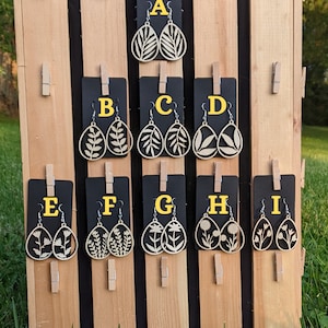 May include: Nine pairs of wooden earrings displayed on a wooden board. Each pair is labeled with a letter from A to I. The earrings are all different designs featuring leaves and flowers.