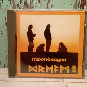 Może przedstawiać: Żółte pudełko na płytę CD z nazwą zespołu "Moondragon" i tekstem "DRMF" w stylizowanym kroju pisma. Pudełko przedstawia sylwetkę czterech osób stojących przed zachodem słońca.