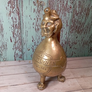 May include: A vintage brass vessel with a devil-like face on top and three clawed feet. The body of the vessel is engraved with the words "ET LUMEN" and "MDLXXV". The vessel has a bulbous shape and a small spout.