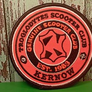 May include: Round patch with the text "TROGLODYTES SCOOTER CLUB GENUINE SCOOTER CLUB EST. 1983 KERNOW" in orange and white on a red and black background. The patch has a black border and a central design.