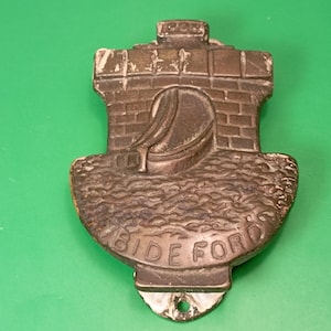 Peut inclure: Un heurtoir de porte de couleur bronze avec un motif de pont. Le heurtoir représente un pont détaillé avec une arche et un bateau. Le mot "BIDEFORD" est inscrit en bas. Le heurtoir a un trou pour le montage.