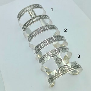 May include: Silver bracelet featuring intricate geometric patterns. The bracelet is crafted from multiple bands stacked on top of each other. The bands are adorned with a variety of geometric designs, including squares, triangles, and circles.