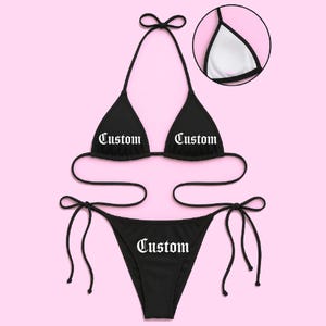 May include: Black bikini set with the word "Custom" printed on the top and bottom. The top is a triangle style with halter straps, and the bottom is a string bikini. The background is a solid pink color.