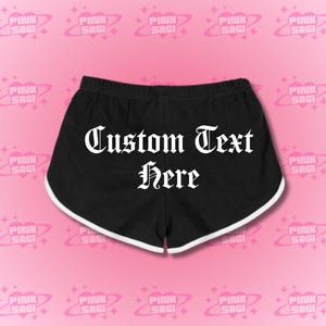 May include: Black athletic shorts with white trim and an elastic waistband. The shorts feature the text "Custom Text Here" in a gothic font. The background is pink with a repeating pattern of the words "Pink Sagi".