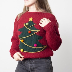 May include: A red knitted sweater featuring a green Christmas tree design with gold garland and colorful pom-poms. A yellow star tops the tree. The sweater has long sleeves and a crew neck.