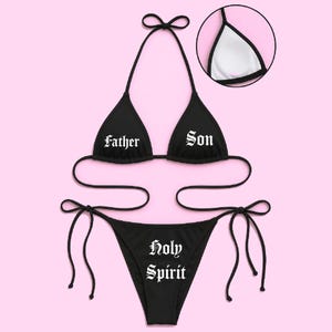 May include: Black bikini set with the words "Father" and "Son" on the top and "Holy Spirit" on the bottom. The bikini top has a halter neck design, and the bottoms are a string style. The background is a light pink color.