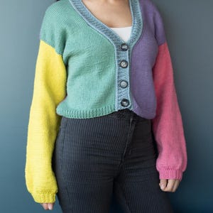 May include: A multi-colored, cropped cardigan sweater. The sweater features a V-neckline and is buttoned down the front. The sleeves are yellow and pink, while the body is green and purple. The sweater is made of a soft, knitted material.