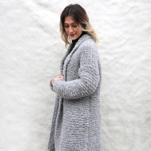 May include: A light grey, long, textured coat with a shawl collar. The coat is made of a soft, fuzzy material and has a relaxed fit. The coat is knee-length and appears warm and comfortable.