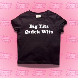 May include: Black t-shirt featuring the white text "Big Tits Quick Wits". The top has a crew neck and short sleeves. The background is pink with a repeating pattern of the words "Pink Sagi" in a circular design.