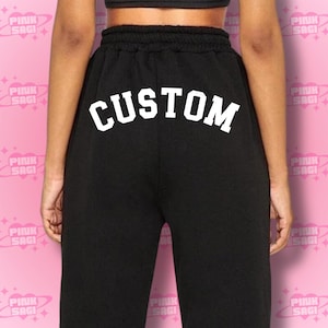May include: Black sweatpants with a white "CUSTOM" text across the back. The pants have a drawstring waist and are made of a soft material. The text is in a bold, block font.