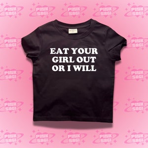 May include: Black t-shirt with white text that reads "EAT YOUR GIRL OUT OR I WILL".