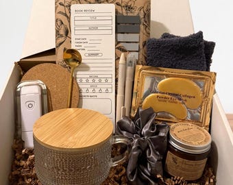 May include: A gift box containing a glass mug with a bamboo lid, a gold spoon, a book review notepad, two pens, a cork coaster, a white portable light, a dark gray fuzzy cloth, gold eye masks, a gray satin scrunchie, and a scented candle in a jar.