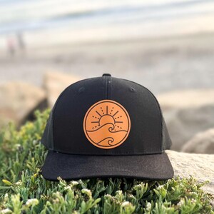 May include: Black trucker hat with a circular orange patch depicting a sun and waves. The hat has a curved peak and a mesh back. The hat is resting on a rock, with greenery in the foreground and a blurred beach scene in the background.
