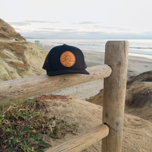 May include: Black trucker hat with a circular patch featuring a sun and wave design in shades of tan and orange. The hat is resting on a wooden fence with a beach and ocean backdrop.
