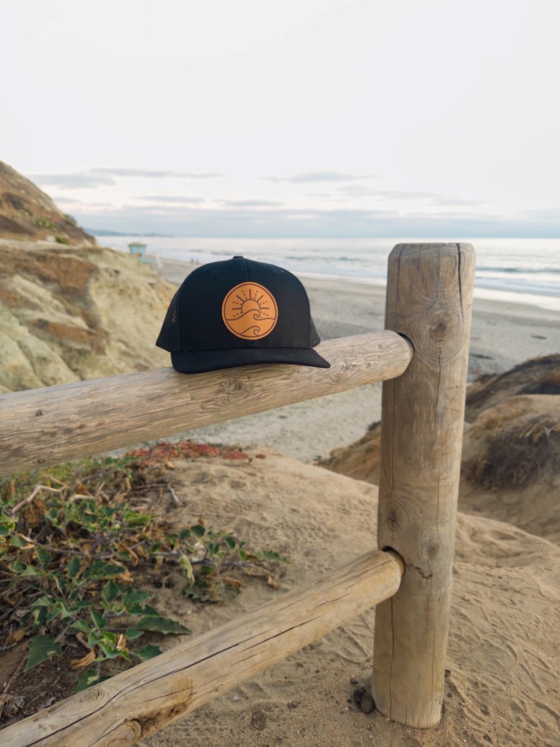 May include: Black trucker hat with a circular patch featuring a sun and wave design in shades of tan and orange. The hat is resting on a wooden fence with a beach and ocean backdrop.
