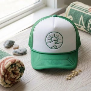 May include: A green and white trucker hat with a circular emblem of a sun and waves. The hat has a green peak and mesh back. Rolled-up towels and small stones are in the background.