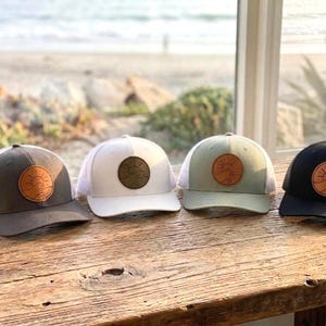 May include: Four trucker hats in a row, each with a different color scheme. The hats have a patch with a sun and wave design. The colors are dark grey, white, sage green, and black. The hats are sitting on a wooden surface.