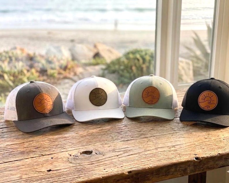 May include: Four trucker hats in a row, each with a different color scheme. The hats have a patch with a sun and wave design. The colors are dark grey, white, sage green, and black. The hats are sitting on a wooden surface.