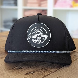 Op de afbeelding: Zwart-witte baseballpet met een witte touwrand en een rond patch met de tekst "Four Boys of Summer" met een zon- en golfgrafiek.
