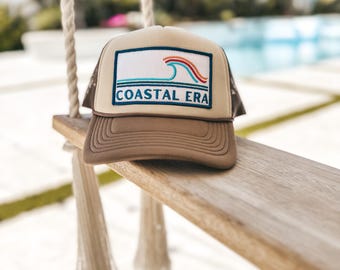 May include: A brown and tan trucker hat with a rectangular patch featuring a wave design in white, red, and blue, and the text "COASTAL ERA". The hat is resting on a wooden swing.