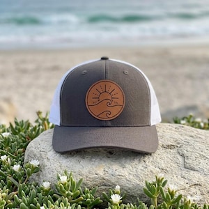 May include: A gray and white trucker hat with a brown leather patch featuring a sun and wave design. The hat has a curved brim and a mesh back. The hat is sitting on a rock with greenery in the foreground and the ocean in the background.