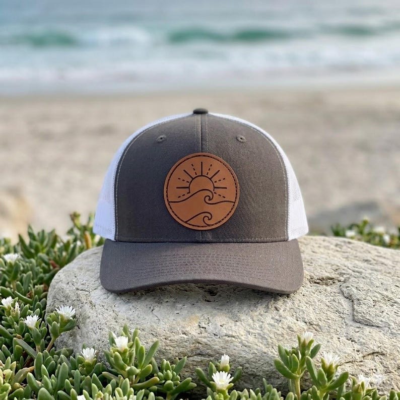 May include: A gray and white trucker hat with a brown leather patch featuring a sun and wave design. The hat has a curved brim and a mesh back. The hat is sitting on a rock with greenery in the foreground and the ocean in the background.
