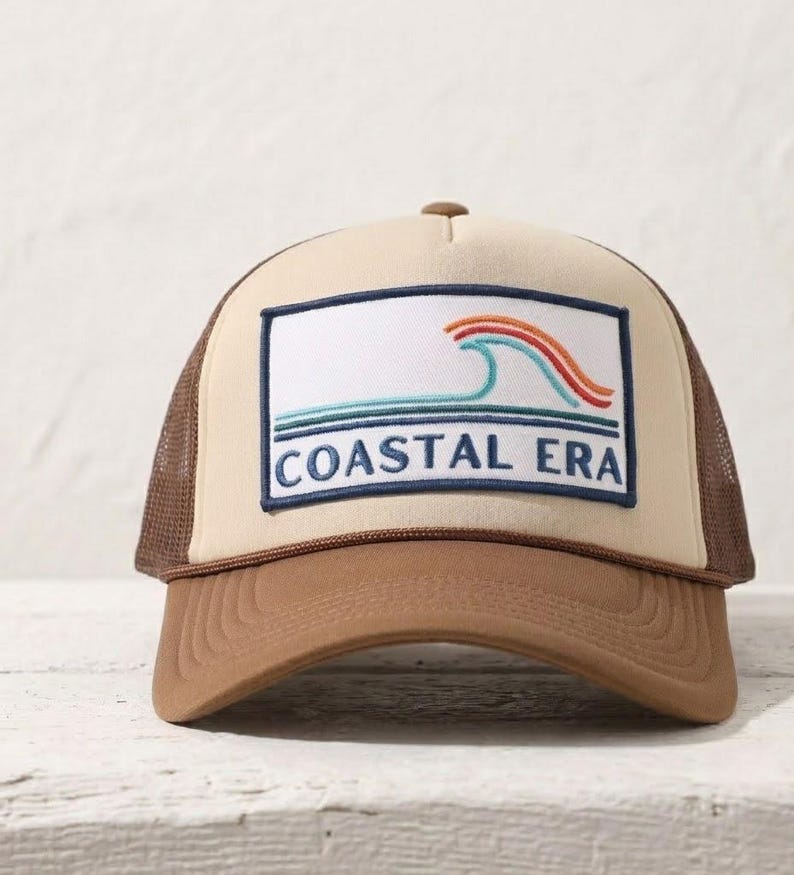 Op de afbeelding: Tan en bruine truckerpet met een rechthoekige patch met een golfontwerp in blauw, oranje en rood, met de tekst "COASTAL ERA" in marineblauw. De pet heeft een bruine klep en een mesh achterkant.