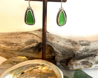 May include: A pair of silver dangle earrings with green sea glass set in a silver bezel. The earrings are displayed on a wooden stand with a shell and sea glass in the background.