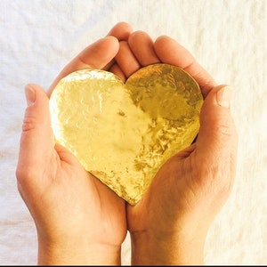 Puede incluir: Un par de manos sosteniendo un plato dorado en forma de corazón. El plato tiene una textura martillada y está hecho de metal.
