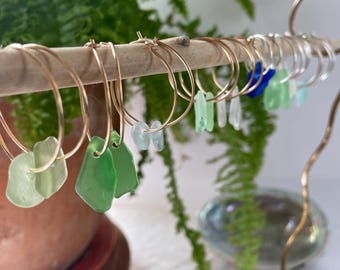 May include: Gold hoop earrings with sea glass charms in shades of green and blue. The earrings are displayed on a wooden stand.