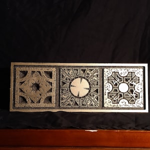 May include: A silver-colored puzzle box with three square panels, each intricately detailed with geometric patterns and symbols. The panels are set within a rectangular frame, creating a cohesive design. The box is displayed against a dark background.