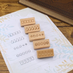 K&ouml;nnte beinhalten: Holzstempel mit verschiedenen Designs, darunter K&auml;stchen, Wassertropfen, Wettersymbole, Gesichter und Gl&auml;ser. Die Stempel sind auf einem gemusterten Papier mit passenden Umrissen angeordnet. Ein Buch ist im Hintergrund zu sehen.