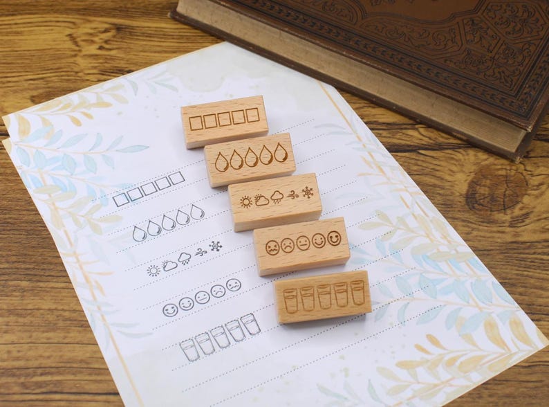 K&ouml;nnte beinhalten: Holzstempel mit verschiedenen Designs, darunter K&auml;stchen, Wassertropfen, Wettersymbole, Gesichter und Gl&auml;ser. Die Stempel sind auf einem gemusterten Papier mit passenden Umrissen angeordnet. Ein Buch ist im Hintergrund zu sehen.
