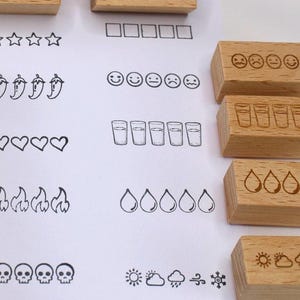 K&ouml;nnte beinhalten: Holzstempel mit verschiedenen Designs, darunter Sterne, Quadrate, Chilischoten, Herzen, Flammen, Totenk&ouml;pfe und Wassergl&auml;ser. Zus&auml;tzliche Stempel zeigen Gesichter mit unterschiedlichen Ausdr&uuml;cken, Wassertropfen und Wettersymbolen. F&uuml;r Bastelarbeiten und Journale.