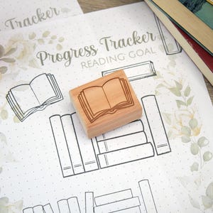 K&ouml;nnte beinhalten: Ein Holzstempel mit einem offenen Buchdesign liegt auf einem "Progress Tracker Reading Goal"-Blatt. Das Blatt hat einen floralen Rand und Buchillustrationen. Ein Stapel B&uuml;cher ist im Hintergrund zu sehen.