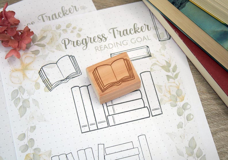 K&ouml;nnte beinhalten: Ein Holzstempel mit einem offenen Buchdesign liegt auf einem "Progress Tracker Reading Goal"-Blatt. Das Blatt hat einen floralen Rand und Buchillustrationen. Ein Stapel B&uuml;cher ist im Hintergrund zu sehen.
