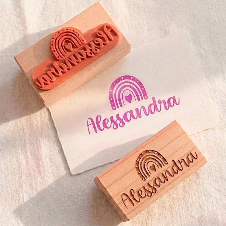 May include: Wooden stamp featuring the name "Alessandra" and a rainbow motif. The stamp is displayed alongside a matching ink print on paper. The stamp is made of wood and has a rubber stamp with the name and rainbow design.
