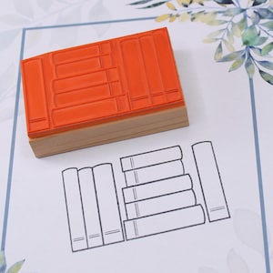K&ouml;nnte beinhalten: Ein orangefarbener Gummistempel mit Holzsockel, der ein B&uuml;cherstapel-Design zeigt. Der Stempel liegt auf wei&szlig;em Papier mit einer gedruckten Umrisszeichnung von B&uuml;chern. Der Hintergrund ist eine Holzoberfl&auml;che.